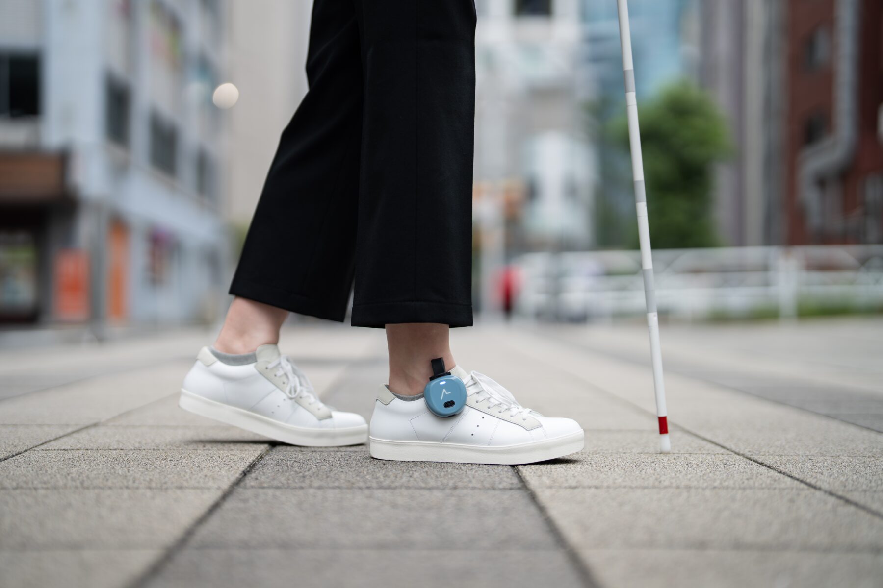 A person walking with a white cane, wearing shoes equipped with the ASHIRASE navigation device.