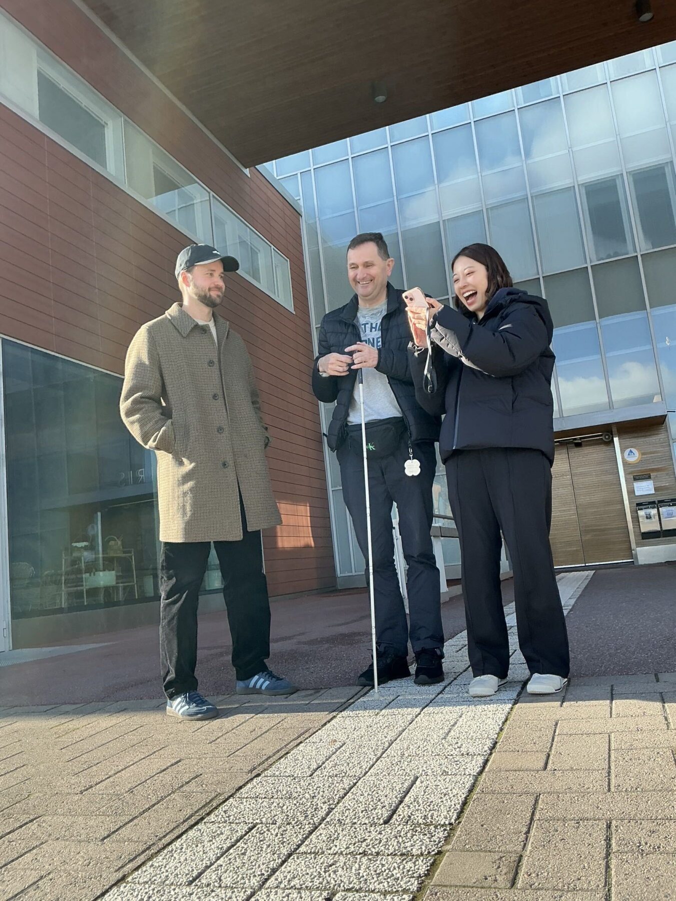 The photo shows three people standing outside the IIRIS Center. The tester is standing in the middle, holding a white cane and smiling broadly. A male staff member from the project stands on the left, and an ASHIRASE staff member stands on the right. The photo captures a warm and cheerful moment during the orientation.